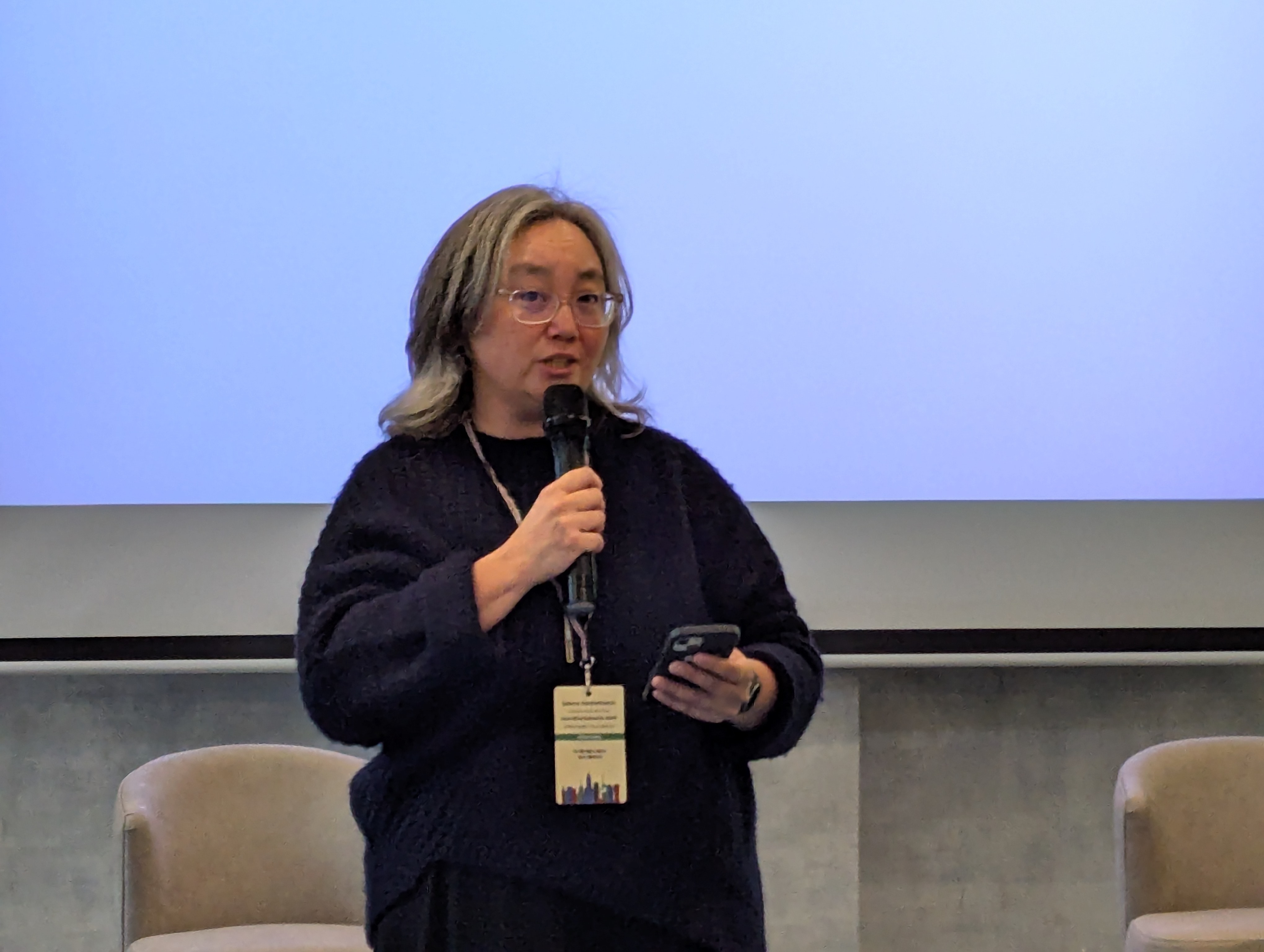 Selena Deckelmann speaking into a handheld microphone, a phone in her other hand, in front of a pale blue projection screen at Wikimania 2025.