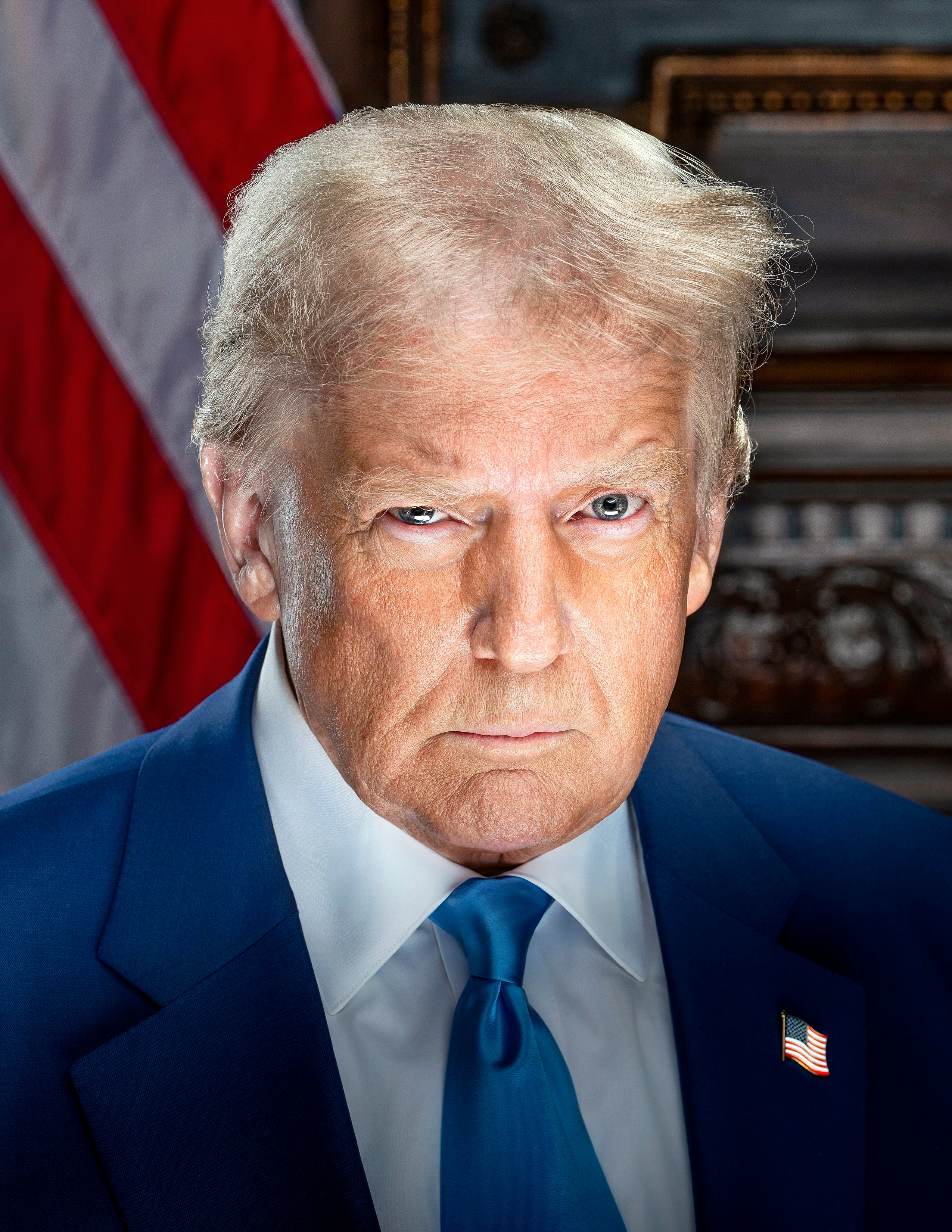 Donald Trump in a dark blue suit, white shirt, blue tie and flag lapel pin, looking at the camera in front of an American flag.