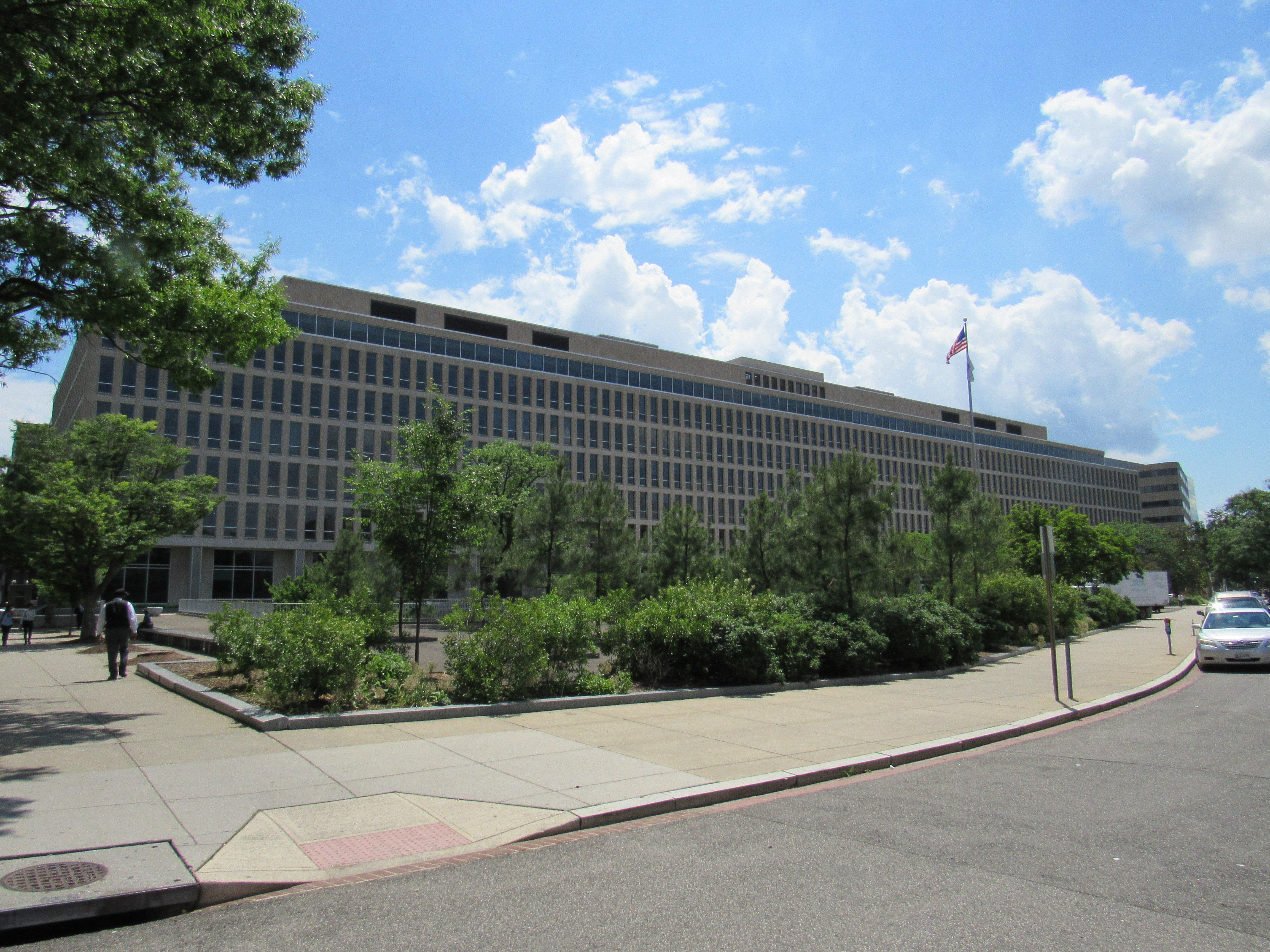 Long, flat-roofed office block of the US Department of Education headquarters in Washington, seen across a landscaped plaza under a blue sky, a US flag on its roof.