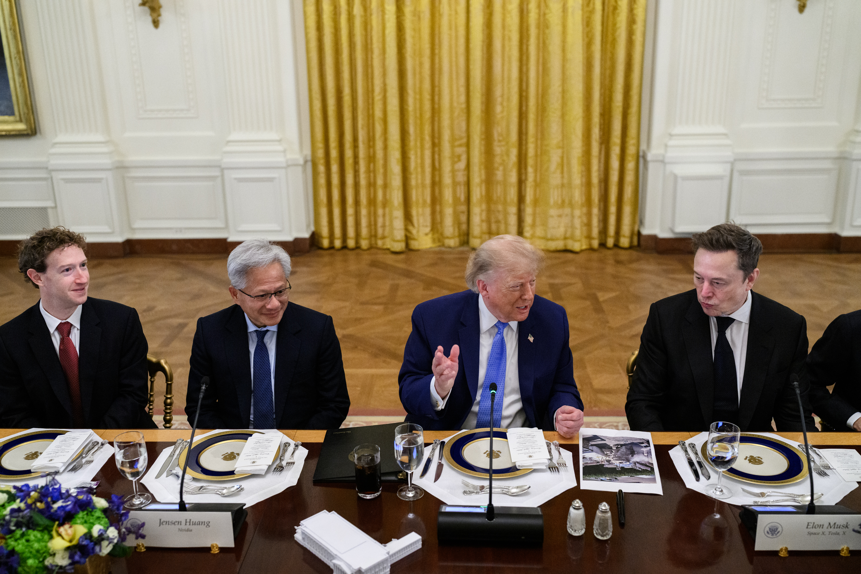 Donald Trump speaking, gesturing with one hand, at a long table in the East Room, seated between Jensen Huang on his left and Elon Musk on his right.