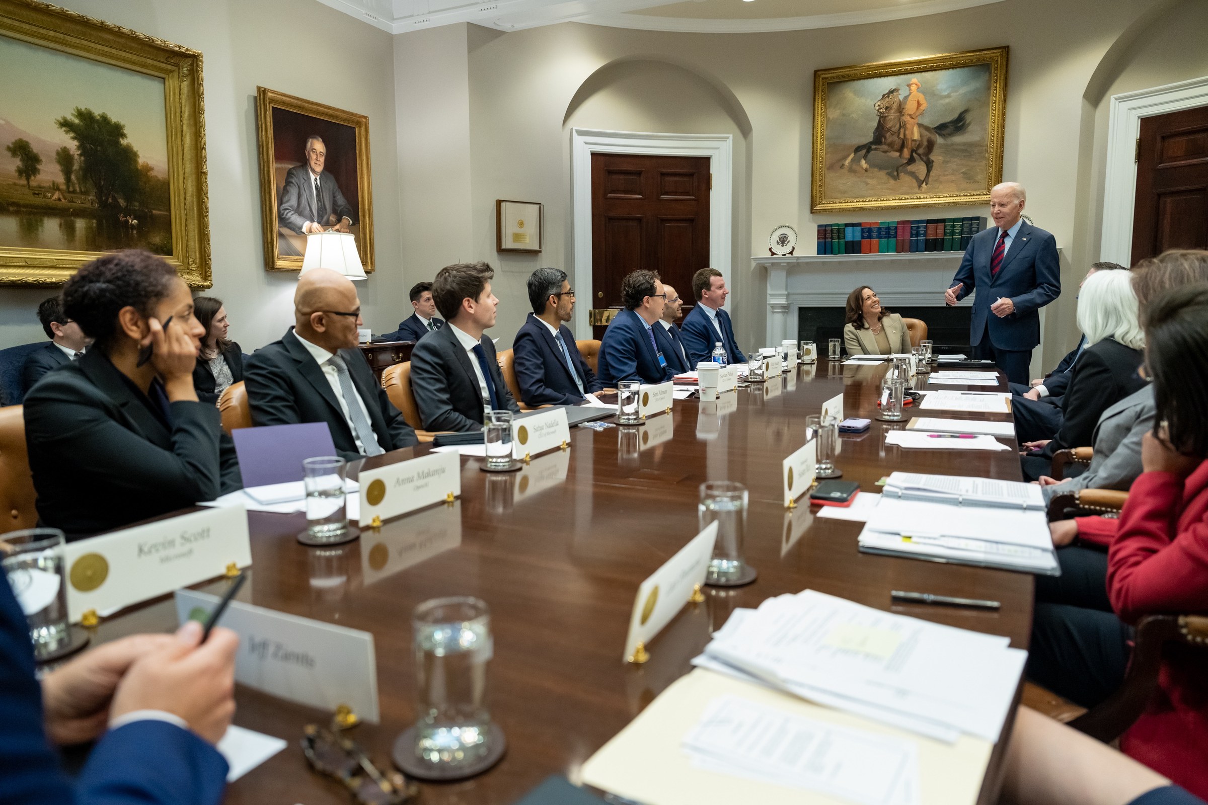 President Biden stands and speaks to AI chief executives seated at a long table; Sam Altman, in a blue tie, listens.