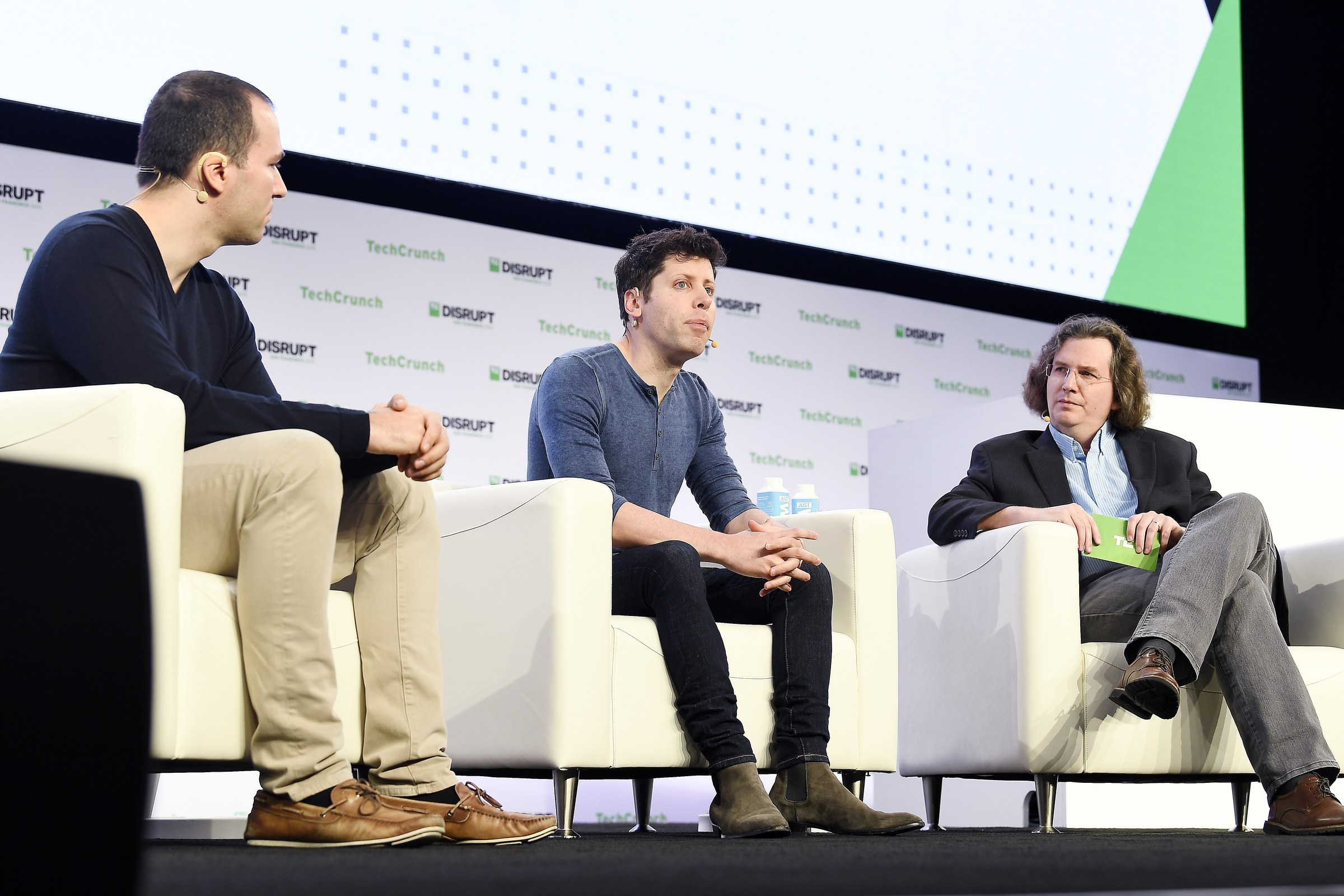 Greg Brockman, Sam Altman and Frederic Lardinois sit in white armchairs on the TechCrunch Disrupt stage.